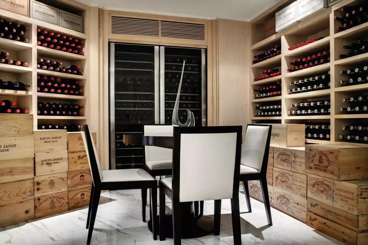 A private cellar features a series of wooden wall shelves and a built-in cooler and at the center of the space stands a small table with four chairs