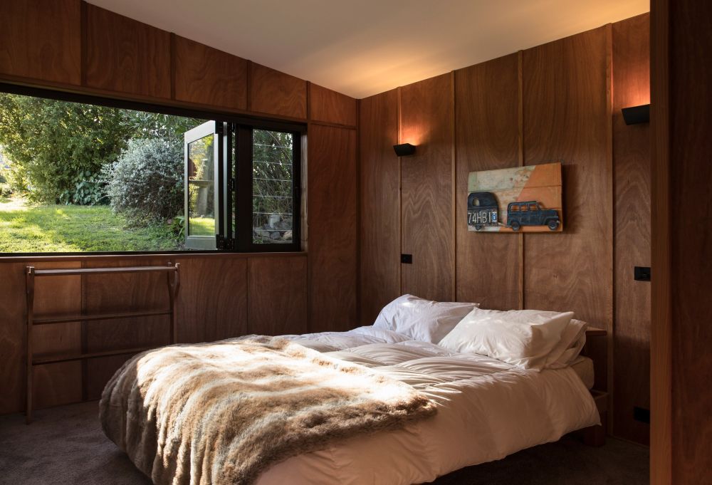This cozy bedroom has walls lined in wood and a long and wide window with peaceful views